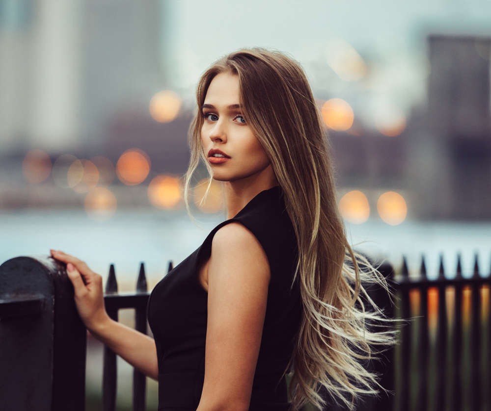 Gorgeous young model woman with perfect blonde hair looking at camera posing in the city wearing black evening dress.